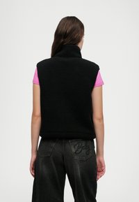 Svart fleecevest med høy krage, kombinert med en rosa t-skjorte og mørk denimbukser med dekorative sømmer på baklommen.