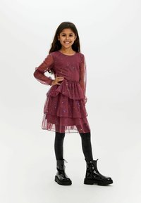 Burgunderfarbene Schichtkleid mit transparenten langen Ärmeln und bunten Paillettenverzierungen, kombiniert mit schwarzen glitzernden Leggings und glänzenden schwarzen Stiefeln.