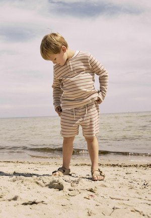 Camicia lunga a maniche con righe beige e pantaloni corti, entrambi con righe rosse. Sandali marroni, in piedi sulla spiaggia vicino all'acqua, guardando in basso.