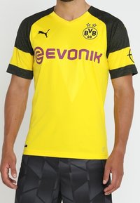 Gul sportströja med svarta texturerade ärmar, med Puma-logotypen, "Evonik" i lila och en klubbemblem med två stjärnor.