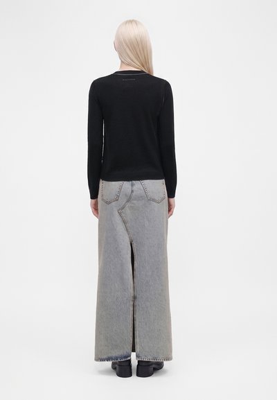 Femme aux longs cheveux blonds portant un haut noir à manches longues et une jupe maxi en denim gris avec une fente dans le dos et des chaussures noires.