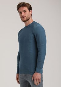 Maglione blu con texture, colletto rotondo, polsini e orlo a coste. Presenta un motivo in rilievo su tutta la superficie e un piccolo logo sul lato inferiore.