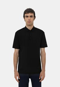 Polo shirt nero a maniche corte con colletto, dotato di un dettaglio con zip sul collo e un logo sulla manica destra. Tessuto morbido.