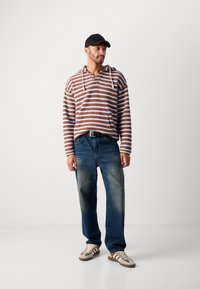 Stripet brun og hvit hettegenser med en frontlomme, parret med blå denimjeans og nøytralt tonede joggesko med striper, samt en svart caps.