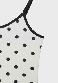Haut blanc sans manches avec fines bretelles noires, bordure noire et pois noirs espacés uniformément sur le tissu.