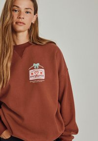 Brun oversized sweatshirt med rund halsudskæring, der har et farverigt grafisk print med teksten "WINTER SPICE" og dekorative elementer.