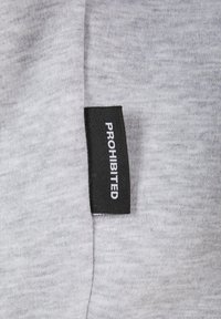 Pilkas audinys su švelnia tekstūra, ant kurio yra juoda etiketė su užrašu "PROHIBITUOTA" baltu šriftu. Etiketė yra šalia audinio krašto.