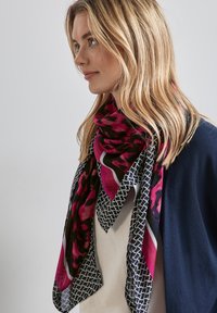 Ampio foulard floreale nero e rosa con bordatura geometrica, realizzato in un tessuto leggero, che si drappeggia attorno al collo, caratterizzato da una superficie liscia.
