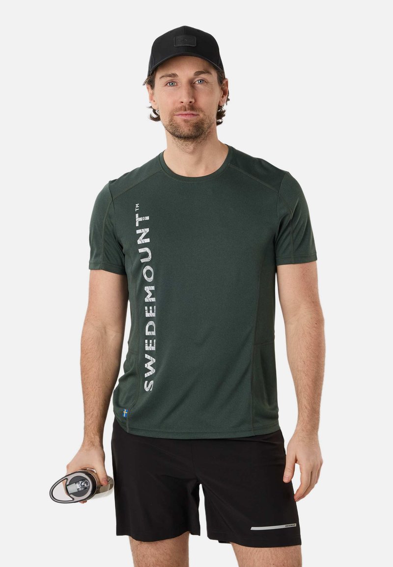 Homme portant un t-shirt Swedemount vert foncé, un short noir et une casquette noire, tenant une bouteille d'eau dans sa main droite.