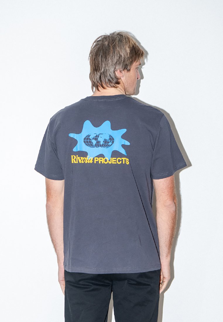 Άνδρας που κοιτάζει προς τα πίσω, φοράει μπλουζάκι σκούρου γκρι με μπλε αφηρημένο σχήμα και το κείμενο "Rivvia Projects" στην πλάτη, μαύρο παντελόνι, στέκεται μπροστά σε λευκό τοίχο.