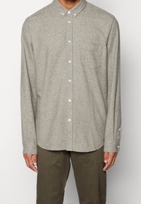 Hellgraues Hemd aus strukturiertem Stoff mit spitzem Kragen, Brusttasche und geknöpften Manschetten, kombiniert mit olivgrünen Hosen.