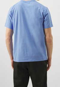 Camiseta de algodón azul claro con mangas cortas, cuello redondo y textura suave, combinada con pantalones negros, vista desde atrás.