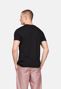 Svart kortermet t-skjorte med rund hals, iført med røde og hvite stripete bukser, sett bakfra. Enkelt design, glatt tekstur.