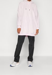 Hoodie oversize rosa in morbido tessuto testurizzato con cappuccio regolabile, abbinato a pantaloni neri a sigaretta e scarpe da ginnastica chiare.