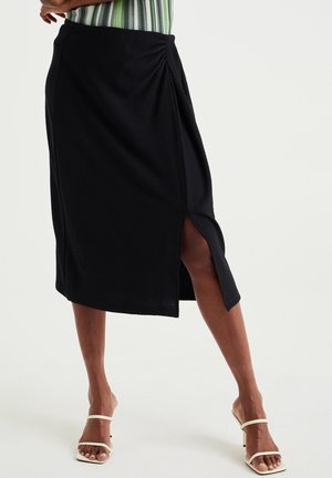 Jupe midi noire avec une fente sur le côté, portée avec des sandales à talons blanches et un haut rayé vert visible à la taille.