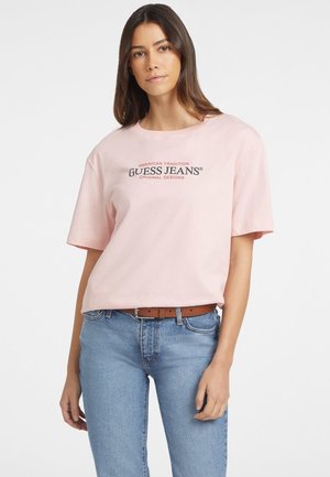 T-shirt en coton rose clair avec des manches courtes et un col rond, portant en son centre le texte noir "GUESS JEANS", associé à un jean bleu.