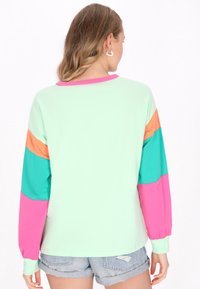 Maglietta a maniche lunghe di colore verde menta con maniche a blocchi di colore in teal, arancione e rosa. Materiale morbido con vestibilità rilassata e colletto a coste.