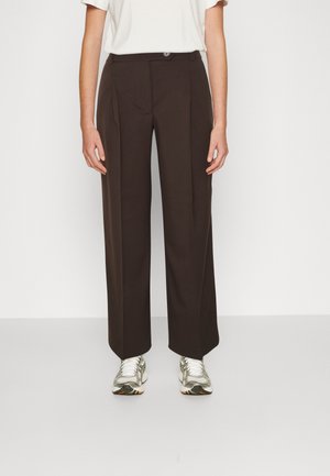 Osoba w prostych, ciemnobrązowych spodniach, białych trampkach i białej koszulce z krótkim rękawem, stojąca na jednolitym tle.