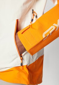Colorful jacket featuring a cream body with orange accents, zipped pockets, and a soft textured sleeve with reflective branding.