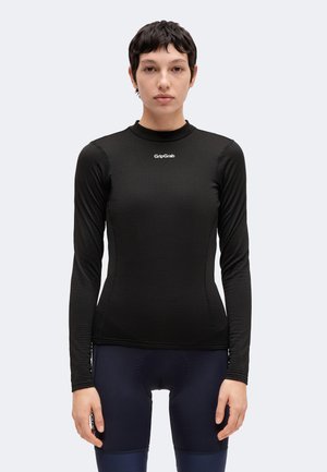 Personne aux cheveux courts et foncés portant un maillot GripGrab noir à manches longues et un cuissard de cyclisme foncé, debout devant un fond uni.