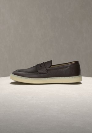 Mørkebrun læderloafer med hvid gummisål, med en penny-rem og tekstureret finish, vist på en lys overflade mod en neutral baggrund.