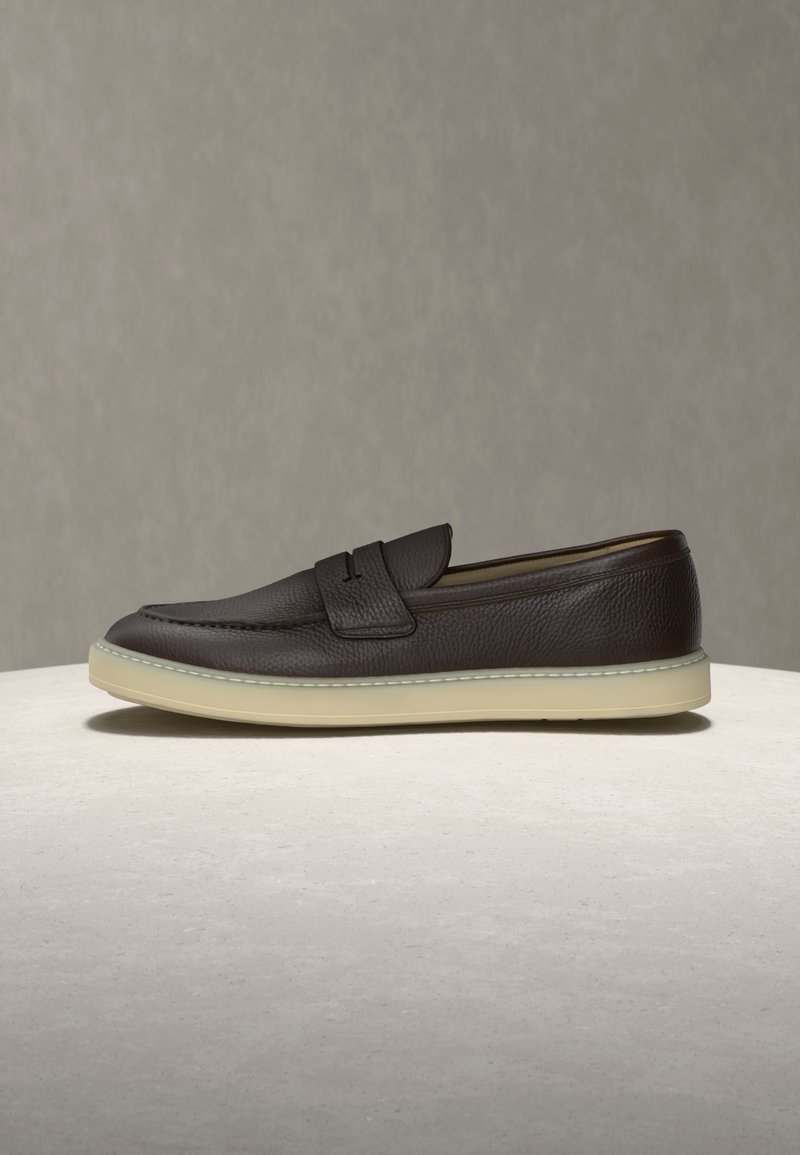 Σκούρο καφέ δερμάτινο μοκασίνι με λευκή λαστιχένια σόλα, με λουράκι penny και υφή, τοποθετημένο σε ανοιχτή επιφάνεια με ουδέτερο φόντο.