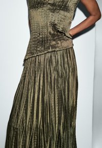 Abito plissettato verde oliva con tessuto testurizzato, caratterizzato da scollo a V e corpetto aderente, che si apre in una lunga gonna a strati.