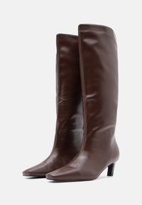 Καφέ δερμάτινες μπότες μέχρι το γόνατο με κομψό σχεδιασμό, μυτερή μύτη και τακούνι 3 ιντσών. Λεία υφή με ελάχιστα μεταλλικά στοιχεία ή σχέδια.