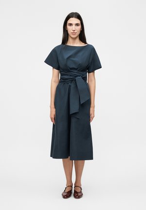 Žena s dlhými tmavými vlasmi stojí oblečená v tmavomodrých midi šatách so širokými krátkymi rukávmi a viazaným opaskom v páse, v kombinácii s tmavočervenými topánkami Mary Jane.