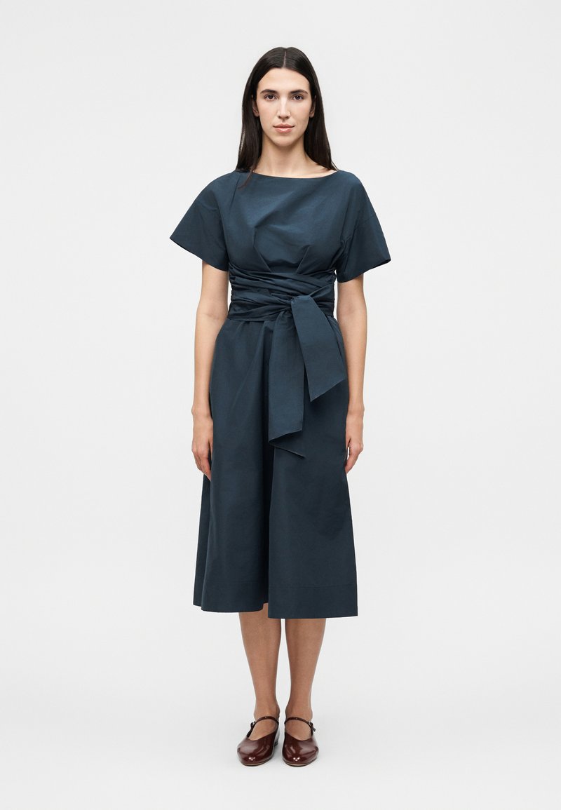 Γυναίκα με μακριά σκούρα μαλλιά στέκεται φορώντας σκούρο μπλε μίντι φόρεμα με φαρδιά κοντά μανίκια και δεμένη ζώνη στη μέση, συνδυασμένο με σκούρα κόκκινα παπούτσια Mary Jane.