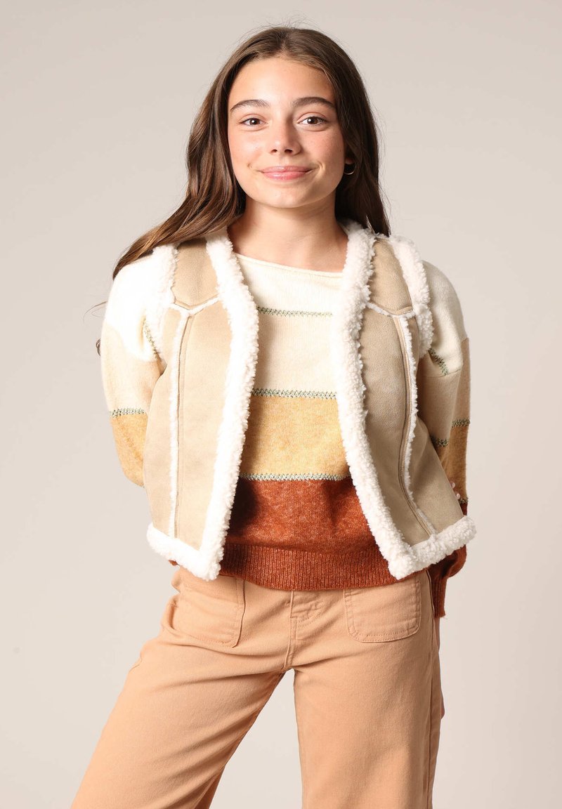 Veste en daim beige avec des bordures en fourrure blanche, superposée à un pull en maille rayé multicolore dans des teintes de crème, orange et marron, accompagnée d'un pantalon beige.