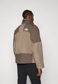 Mężczyzna w brązowej i beżowej kurtce North Face z wzorzystymi panelami, stojący tyłem z głową zwróconą w prawo na jednolitym tle.