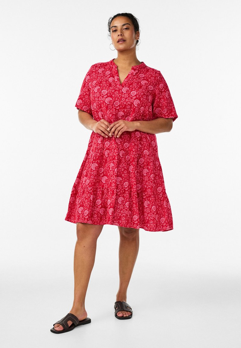 Kvinde står med hænderne foldet, iført en rød knælængde kjole med blomsterprint og sorte flade sandaler med åbne tæer mod en hvid baggrund.