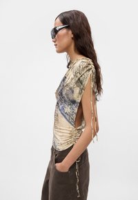 Korte mouwen top in beige en blauw met een gerimpeld ontwerp en uitsnijdingen aan de zijkant. Heeft striksluitingen op de schouders, gemaakt van lichtgewicht materiaal.