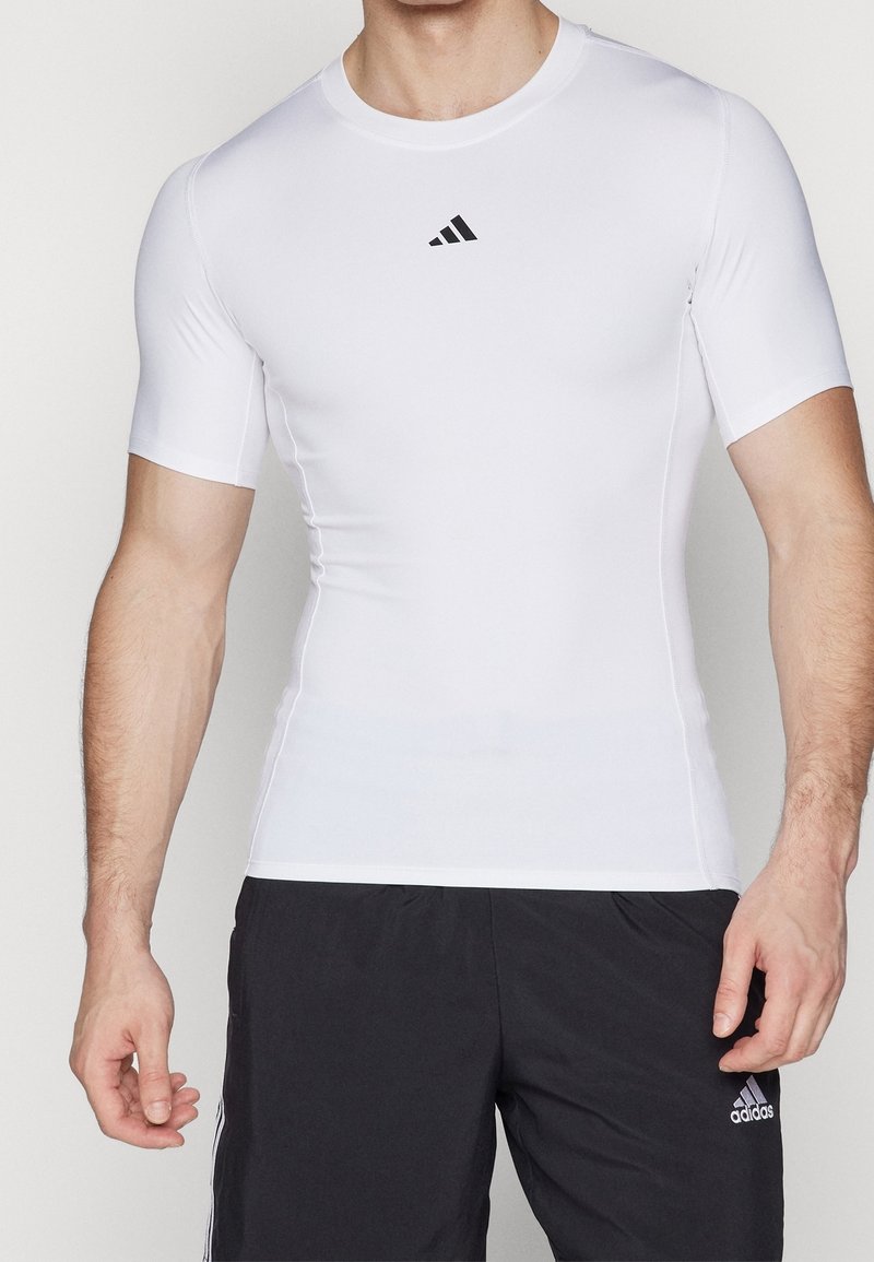 Vit kortärmad idrottströja gjord av glatt, stretchigt tyg. Har en svart Adidas-logotyp på bröstet och en åtsittande design.