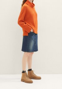 Oranžový svetr s ribbed vzorem a vysokým límcem, v kombinaci s džínovou sukní ke kolenům. Hnědé kotníkové boty na šněrování s texturovaným povrchem.