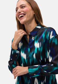 Camicia di raso con un motivo blu, verde e bianco su sfondo nero, dotata di colletto classico e bottoni decorativi sui polsini.