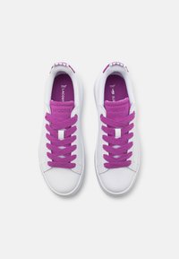 Weiße Sneaker mit lila Schnürsenkeln und Innenfutter. Sie verfügen über eine flache Gummisohle und eine gepolsterte Zunge mit einem Logodetail an der Ferse.
