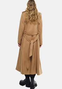 Kamel-farbener, langer Trenchcoat mit Bindegürtel, großem Kragen und seitlichen Schlitzen. Kombiniert mit klobigen schwarzen Stiefeln und gerippten Bündchen.