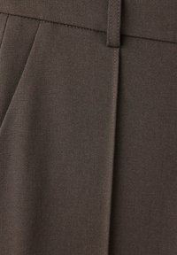 Gros plan d’un pantalon en tissu marron montrant la ceinture, les passants, les plis et les surpiqûres détaillées.