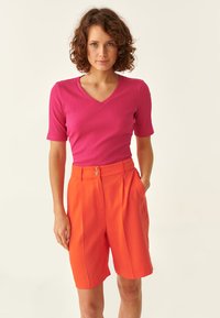 Top rosa a costine con scollo a V aderente abbinato a pantaloni corti arancioni a vita alta con due bottoni frontali. Lo sfondo semplice mette in risalto l'outfit.