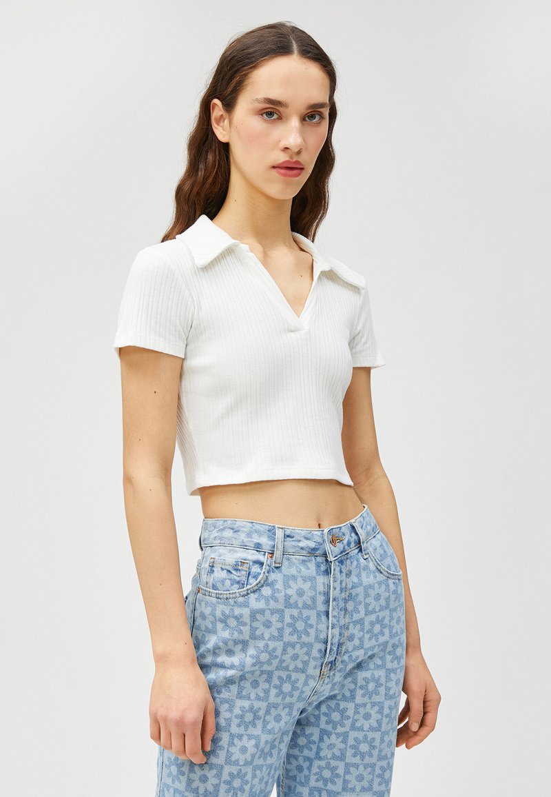 Weißes geripptes Croptop mit Kragen und kurzen Ärmeln, kombiniert mit hellblauen, floral gemusterten Hochbundjeans.