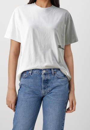 Kobieta w gładkiej białej koszulce z krótkim rękawem, z małym czarnym napisem "KARL JEANFELD" i w wysokich niebieskich dżinsach na neutralnym tle.