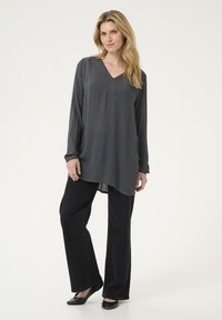Tunic grigia a maniche lunghe con scollo a V, caratterizzata da un tessuto texture e un orlo allungato, abbinata a pantaloni neri a gamba larga e ballerine nere.