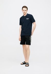 Jeune homme debout portant un polo noir Moschino, un short noir avec un texte blanc sur le côté, et des claquettes noires Moschino sur un fond uni.