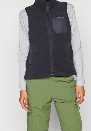 Personne portant un gilet polaire noir zippé sur des manches longues gris clair et un pantalon cargo vert olive, mains détendues le long du corps.