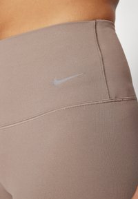 Hnědé legíny s vysokým pasem vyrobené z elastického materiálu, které mají jemné šedé logo Nike a ploché švy pro pohodlí.