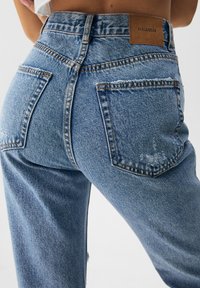 Jeans de mezclilla azul con bolsillos traseros, pequeños detalles desgastados y una etiqueta marrón "PULL&BEAR" en la cintura, llevados por una persona de pie.