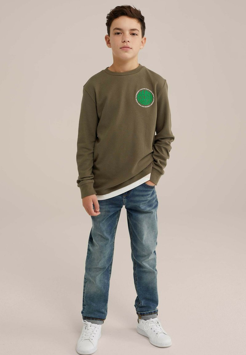 Camicia a maniche lunghe verde oliva con tessuto testurizzato, logo ricamato rotondo, indossata sopra una camicia bianca, abbinata a jeans azzurri e sneakers bianche.