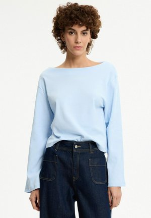 Mujer con cabello corto y rizado lleva una camiseta azul claro de manga larga y vaqueros oscuros de cintura alta, de pie sobre un fondo liso.